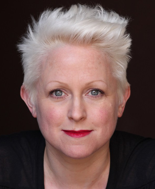 A photograph of Naomi Paxton. We can she her head and shoulders. She has short white-blonde hair, blue eyes, and is wearing red lipstick. She's wearing a black shirt and the background is dark, giving a dramatic image.