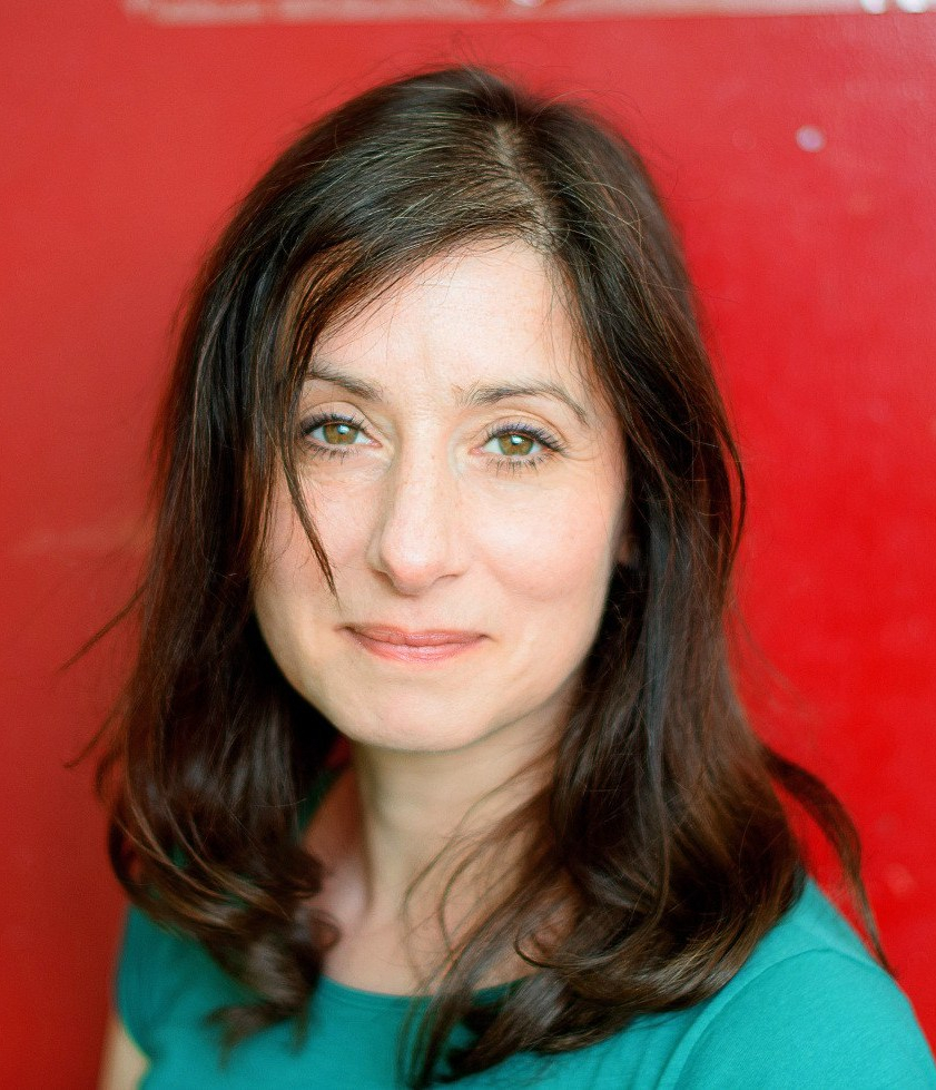 A photograph of Rachel Souhami. We can see her head and shoulders. She has shoulder-length brown hair, is wearing a green t-shirt and is standing in front of a red background.